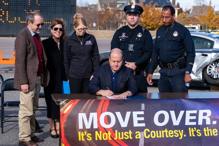 Government officials, safety advocates, law enforcement and CDOT standing around Governor Polis while he signs the proclamation.