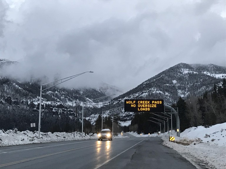 A car drives on a road with a Variable Message Sign overhead that reads "Wolf Creek Pass No Oversize Loads"