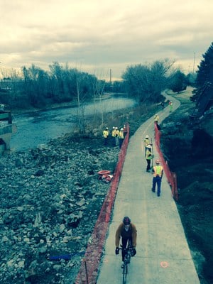 A cyclist using a concrete sidewalk through a construction site with several construction workers on the sidewalk.