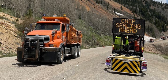 An orange dump truck on a gravel road next to a electonic message board that says, "Chip Sealing Ahead."