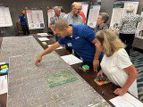 A man wearing a blue polo shirt pointing at a location on a map. A woman wearing a white blouse observing.