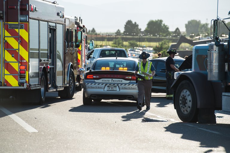 Colorado State Patrol officer standing near a CSP vehicle, firetruck and 18-wheeler at the scene of a car crash.