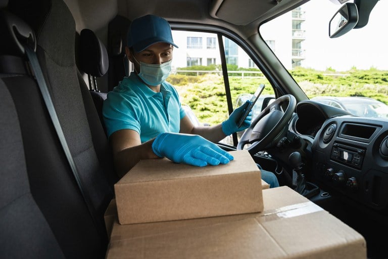 Delivery driver wearing blue shirt, hat and gloves. The person is sitting in a delivery van, holding a cellphone and looking at packages.