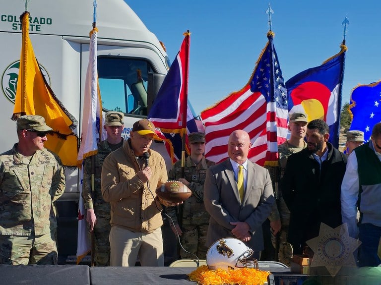 Colorado and Wyoming law enforcement representatives holding footballs and standing in front of flags and a white Colorado State University truck. 