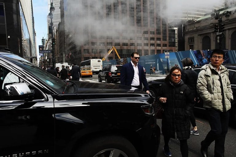 Pedestrians walking in front of a tall black SUV on a busy city street.