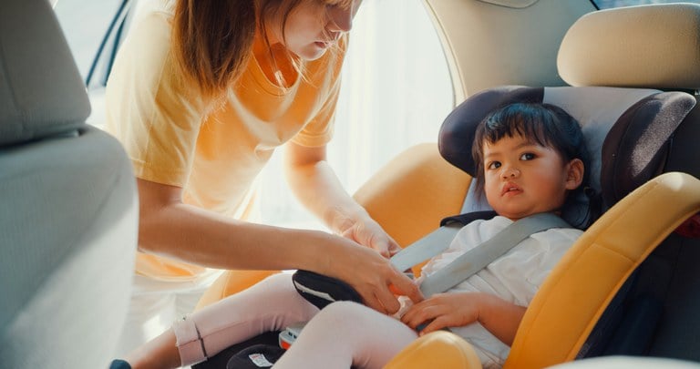 Woman wearing yellow shirt and buckling a child into a car seat in the back of a vehicle.