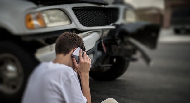 Young man talking on a cellphone and sitting on a curb near a crashed white pickup truck.