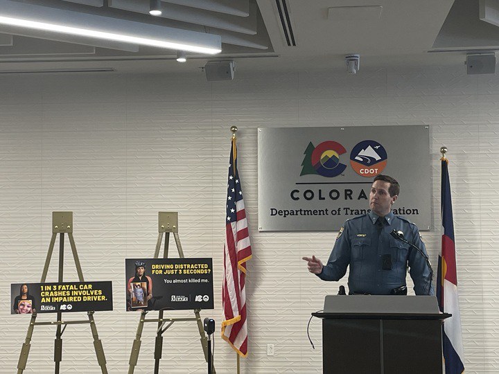 Col. Matthew Packard from Colorado State Patrol addressing last year's (2023) traffic fatalities from behind a podium. He is accompanied by two Shift into Safe posters and the Colorado Department of Transportation placard. 