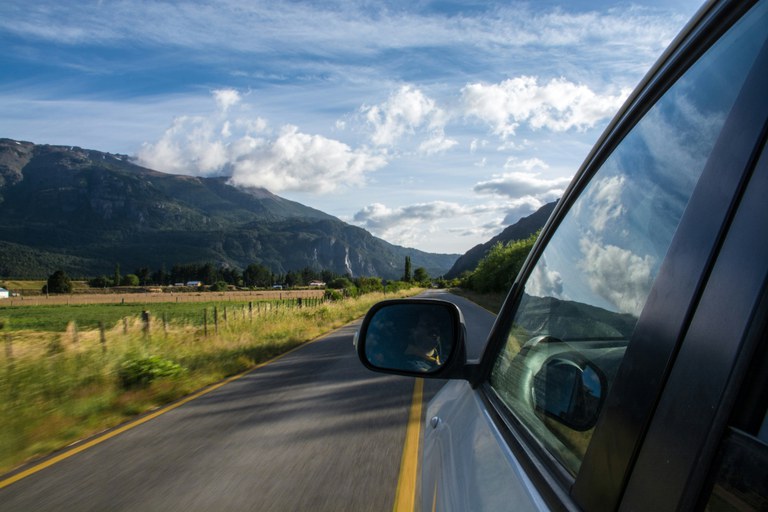 Left side view of a grey car on a mountain road with a grassy field to the left, mountains in front of the car and partly cloudy blue skies.