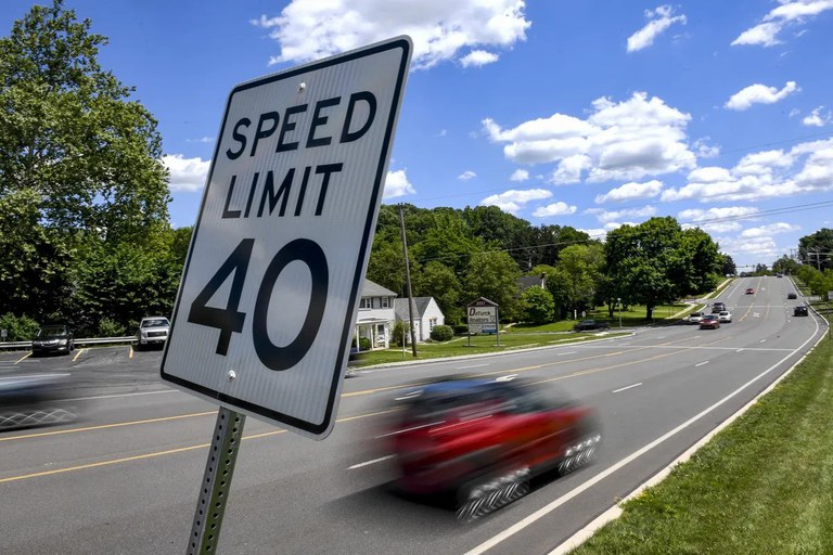 Forty miles per hour speed limit sign beside a busy road with lush green trees and partly cloudy blue skies. A red car is driving on the road.