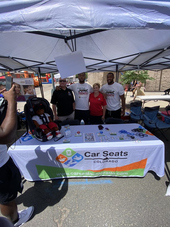 The Car Seats Colorado team standing behind a table and under a white tent at the Juneteenth Festival in Denver. The table is covered with a white tablecloth that displays the Car Seats Colorado logo.