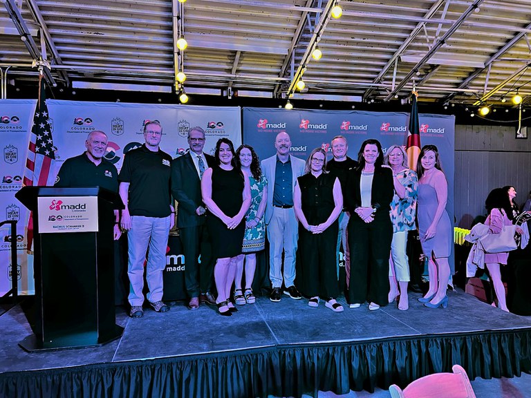 The LEL team and staff members from the Highway Safety Office standing on a stage at the 2024 MADD/CDOT Law Enforcement Champion Awards in Denver. The podium and backdrop have the Colorado Department of Transportation logo and Mothers Against Drunk Driving logo.