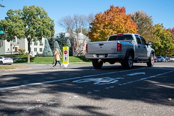 Silver pickup truck stopped in front of a pedestrian crosswalk. A pedestrian is walking in the cross walk and there are houses and colorful large trees along the road.