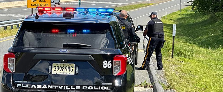 Black Pleasantville Police SUV with flashing lights stopped behind a black car on a highway feeder road. Two Pleasantville police officers are standing beside the car and talking to the motorist who was pulled over for a traffic stop as part of the EPJETS research project.