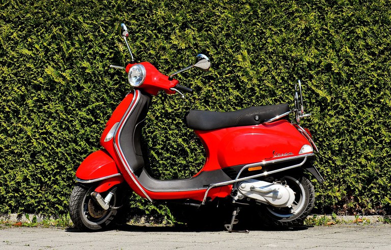 Red Vespa moped on a paved sidewalk in front of a green grass wall.