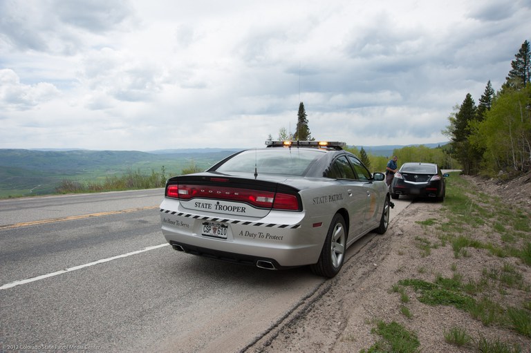 Colorado State Patrol vehicle stopped behind a black sedan on a mountain road with trees in the background and cloudy skies. A Colorado State Patrol trooper is standing on the driver's side of the black sedan. 