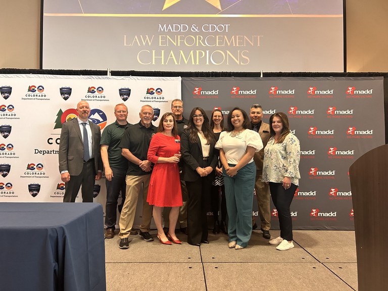 CDOT's Law Enforcement Liaison team standing on a stage at the 2024 MADD/CDOT Law Enforcement Champion Awards in Grand Junction, Colorado. Behind the group of people is a backdrop with CDOT's logo, and backdrop with MADD's logo and a slideshow display with text that reads, "MADD & CDOT Law Enforcement Champions."