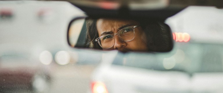 Person with long black hair and clear rectangular glasses looking into their rear view mirror on a busy road.