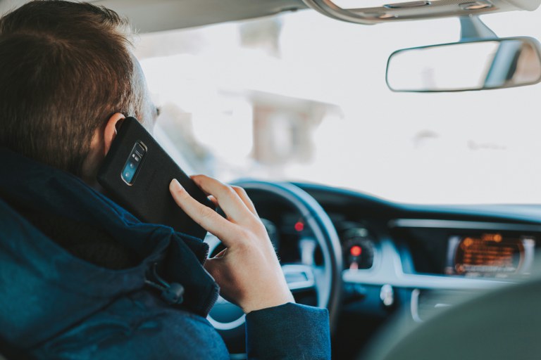 Person with short brown hair holding a black cell phone up to their ear while driving a vehicle.