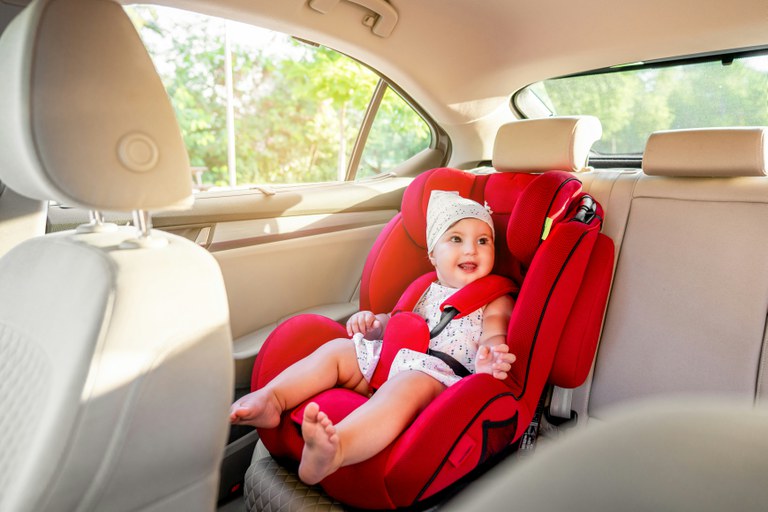 Toddler in a white outfit sitting in a red car seat in the back seat of a vehicle. 