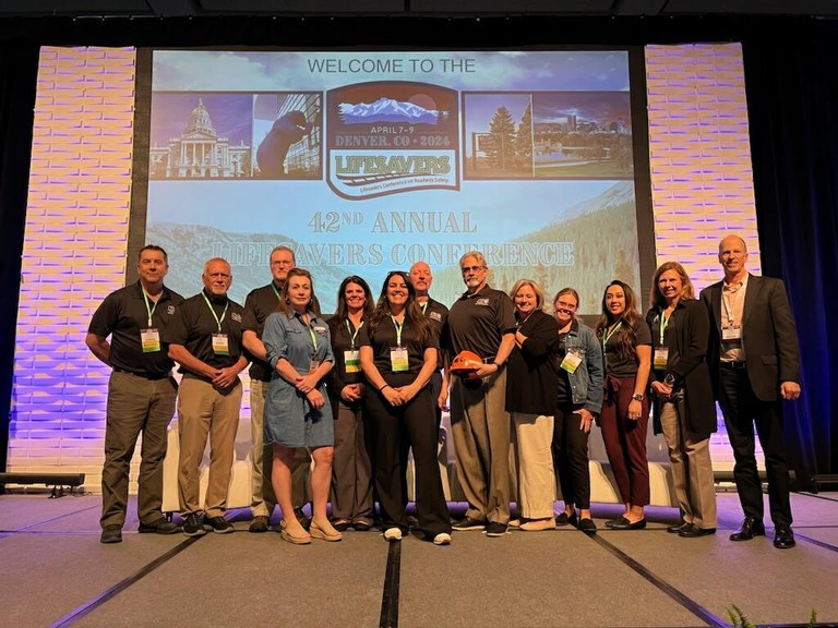 CDOT's Law Enforcement Liaison team standing on a stage at the 42nd Annual 2024 Lifesavers Conference on Roadway Safety the Denver Convention Center. Behind the group of people is a slideshow display with text that reads, "welcome to the 42nd Annual 2024 Lifesavers Conference."