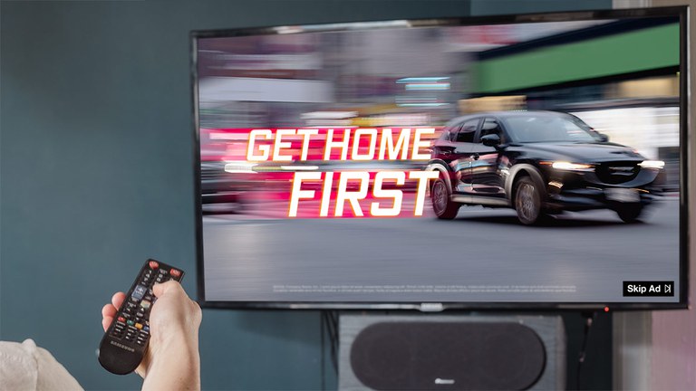 A person holding a remote up to a TV. On the TV is a commercial for a SUV with text that reads, "get home first."