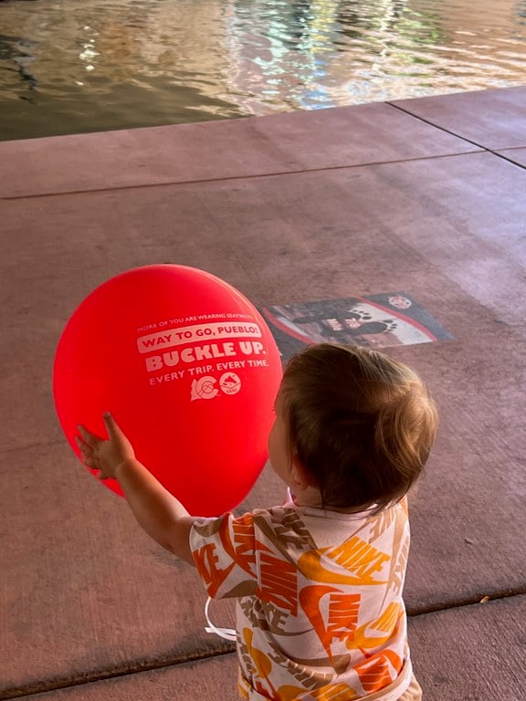 Toddler holding a red balloon on the Historic Arkansas Riverwalk of Pueblo. Text on the balloon reads, "Way to go, Pueblo! Buckle up. Every trip. Every time."