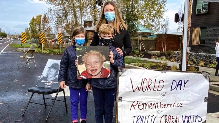 A woman and two children holding up a picture of a toddler during the World Day of Remembrance for Road Traffic Victims event. A poster next to them reads, "World Day of Remembrance for Traffic Crash Victims."