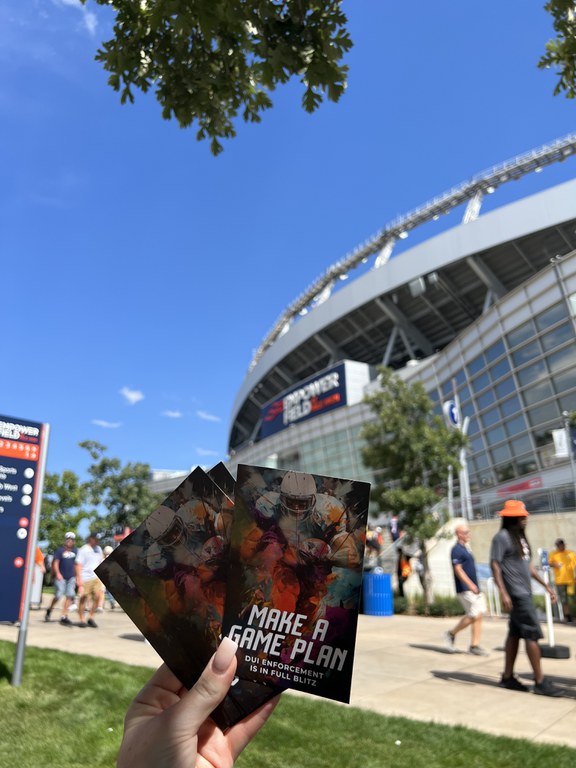 Person holding up Uber passes in front of Empower Field before the Denver Broncos’ home opener with people walking in the background. The passes have a colorful graphic with an illustration of a football player running with football. On graphic copy reads "Make a game plan. DUI enforcement is in full blitz."