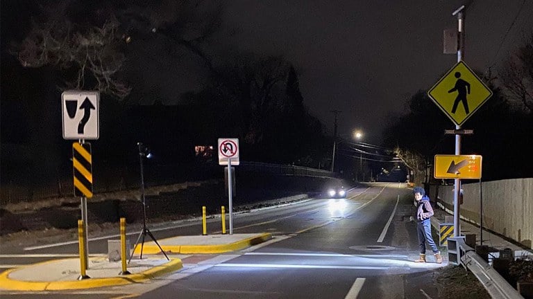 Pedestrian entering a crosswalk at night with a car driving toward the crosswalk. The crosswalk has an illuminator and a rectangular rapid flashing beacon to make the pedestrian more visible.