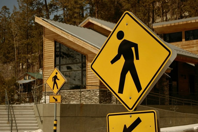 Yellow pedestrian crossing signs near brown wooden building and trees.