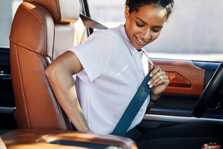 Woman wearing white shirt buckling seat belt in the driver's seat of a vehicle.