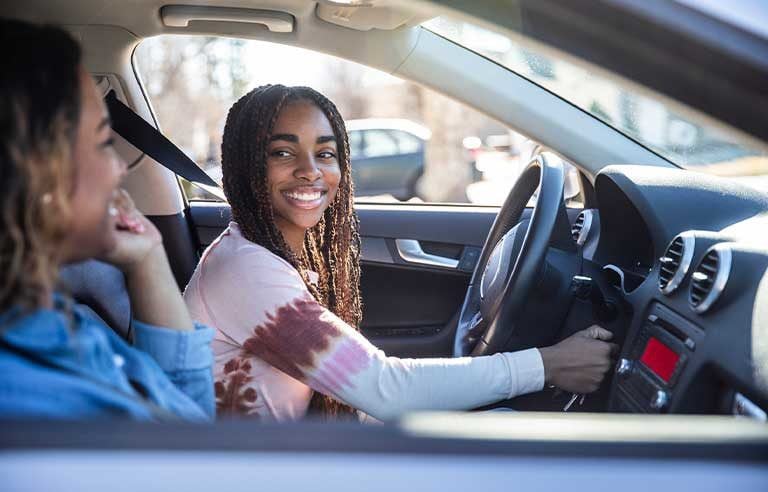 Young adult wearing pink shirt sitting in the driver's seat of a car and looking over at an older adult in the passenger's seat.