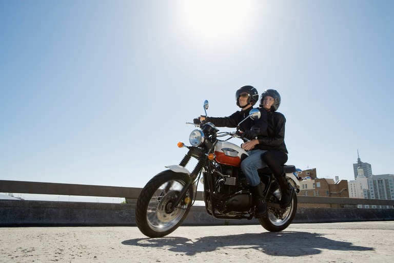 Couple riding motorcycle on city street with buildings and blue sky in the background.  