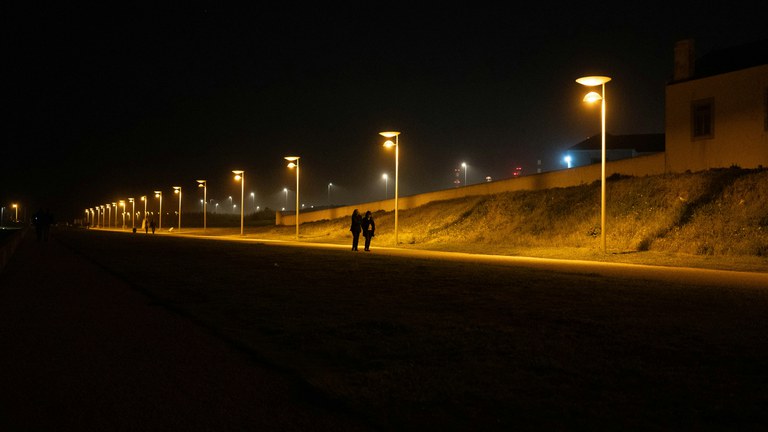 People walking along a road at night with yellow streetlights illuminating the sidewalk. 