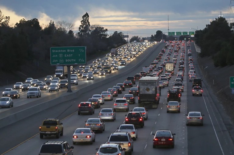 Heavy traffic on a highway at dusk with tall green trees lining the road. 