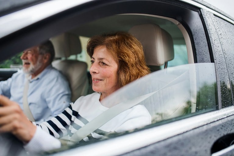 Older woman wearing white shirt driving a black car with an older man wearing blue shirt in the passenger seat.