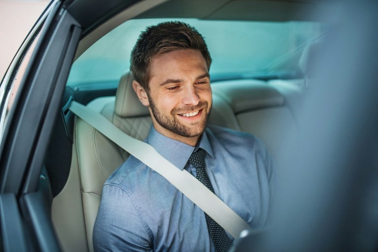 Man wearing blue button-up shirt sitting in the backseat of a vehicle with a seat belt on.