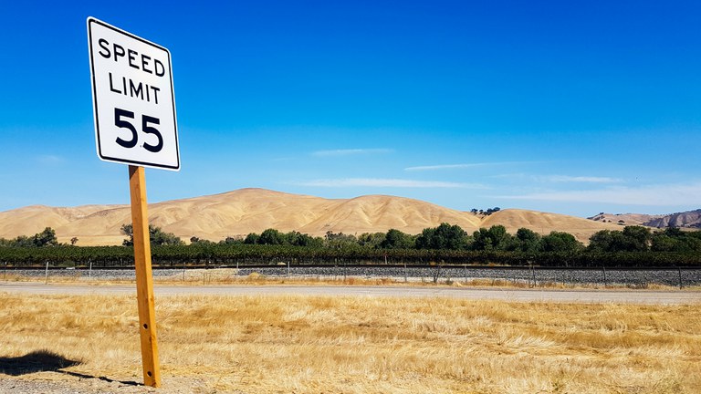 Fifty five miles per hour speed limit sign on a rural road with tan hills and a blue sky. 