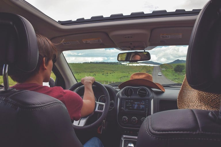 Young man wearing red shirt driving an SUV on a rural road with a cowboy hat on the vehicle's dashboard.