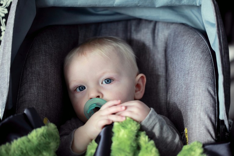 Toddler with blue eyes and blonde hair sitting in a grey car seat and holding a green blanket.